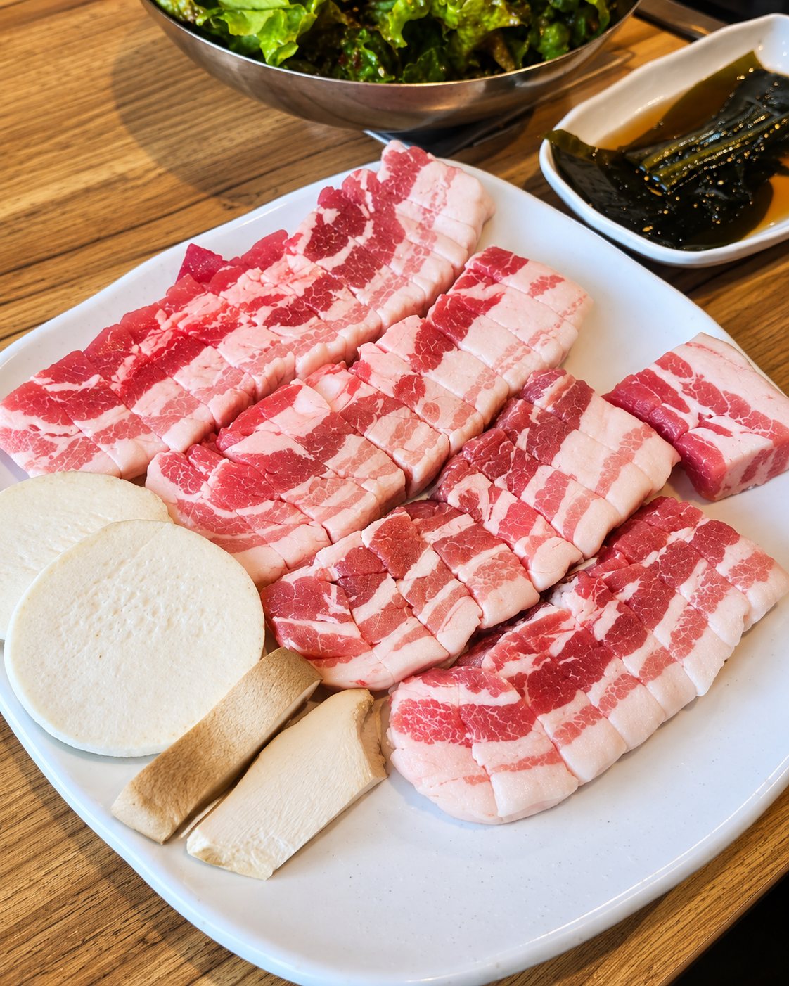 흰 접시에 담은 두툼한 생 삼겹살과 새송이, 곁들이